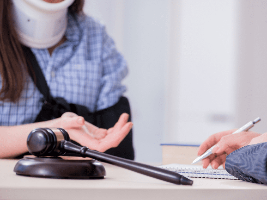 Woman in a hospital gown, neck brace, and arm sling sitting with a lawyer doing paperwork.