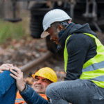 Two men in hardhats and safety vests. One man is on the floor holding his knee in pain.